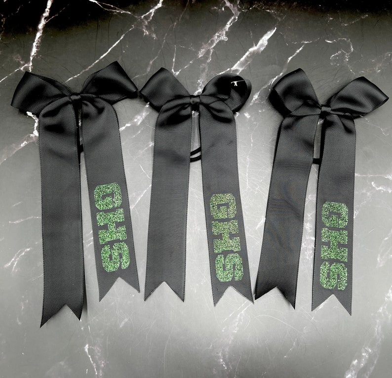 May include: Three black hair bows with long, ribbon tails. Each tail features the green glitter text "GHS". The bows are arranged on a dark, marbled surface.
