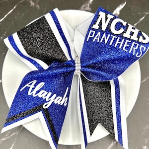May include: A large cheer bow featuring blue and black glitter fabric with white ribbon accents. The bow is centered with a small rhinestone band. The text NCHS PANTHERS is printed in white on the right side, and the name Alayah is printed in white on the left side.