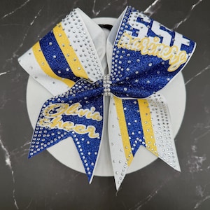 Op de afbeelding: Een grote cheerleading strik met een witte basis, versierd met blauwe en gele glitterstrepen. De strik heeft de tekst "SST Marines" en "Olivia Cheer" in gouden letters, versierd met strass. Een strass sluiting zet het midden van de strik vast.