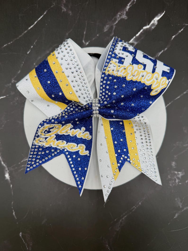 Op de afbeelding: Een grote cheerleading strik met een witte basis, versierd met blauwe en gele glitterstrepen. De strik heeft de tekst "SST Marines" en "Olivia Cheer" in gouden letters, versierd met strass. Een strass sluiting zet het midden van de strik vast.
