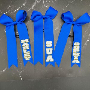 May include: Three blue hair bows with personalized text in gold lettering. The names "MARLEY", "SUA", and "SOPHIA" are displayed on the ribbons. Each bow is tied with a knot and attached to a black elastic band.