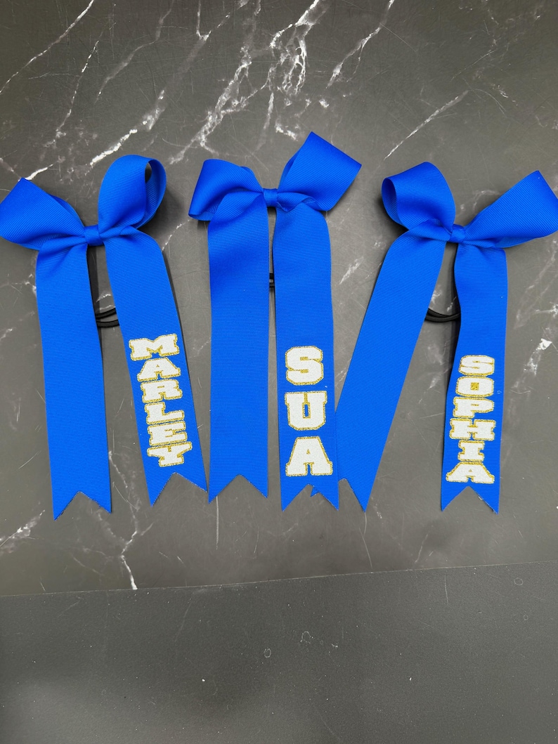 May include: Three blue hair bows with personalized text in gold lettering. The names "MARLEY", "SUA", and "SOPHIA" are displayed on the ribbons. Each bow is tied with a knot and attached to a black elastic band.