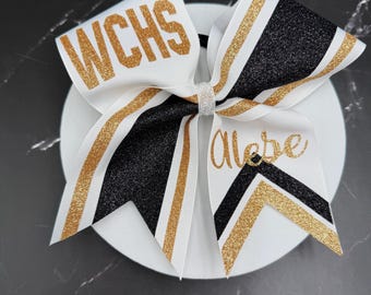 May include: A cheer bow featuring white, black, and gold glitter fabric. The left side displays the text WCHS in gold glitter, while the right side features the name Alebe in gold script. The bow is secured with a silver glitter center knot.