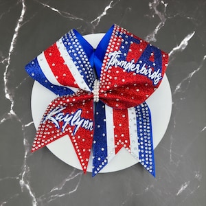 Op de afbeelding: Een rood, wit en blauw cheerleading strik met de naam "Kaylynn" en het woord "Thunderbirds" in witte letters. De strik is versierd met strass en heeft een glinsterende afwerking. Een zilveren strass sluiting zet de strik vast.
