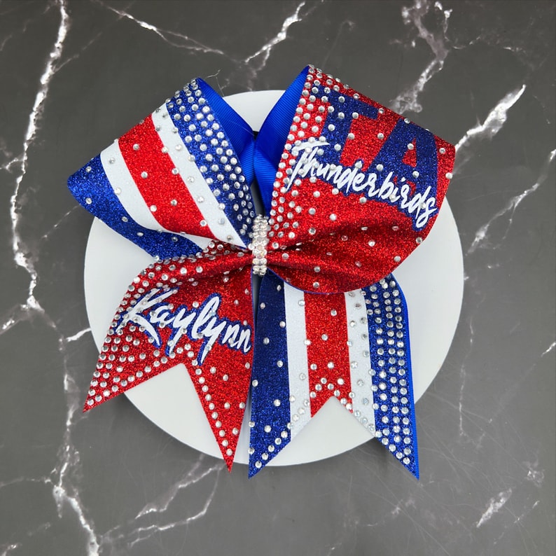 Op de afbeelding: Een rood, wit en blauw cheerleading strik met de naam "Kaylynn" en het woord "Thunderbirds" in witte letters. De strik is versierd met strass en heeft een glinsterende afwerking. Een zilveren strass sluiting zet de strik vast.