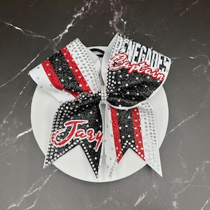 Op de afbeelding: Een cheerleading strik met een wit, zwart en rood ontwerp. De strik heeft de woorden "Renegades Captain" en "Jary" in rode en witte letters. De strik is versierd met strass-steentjes.