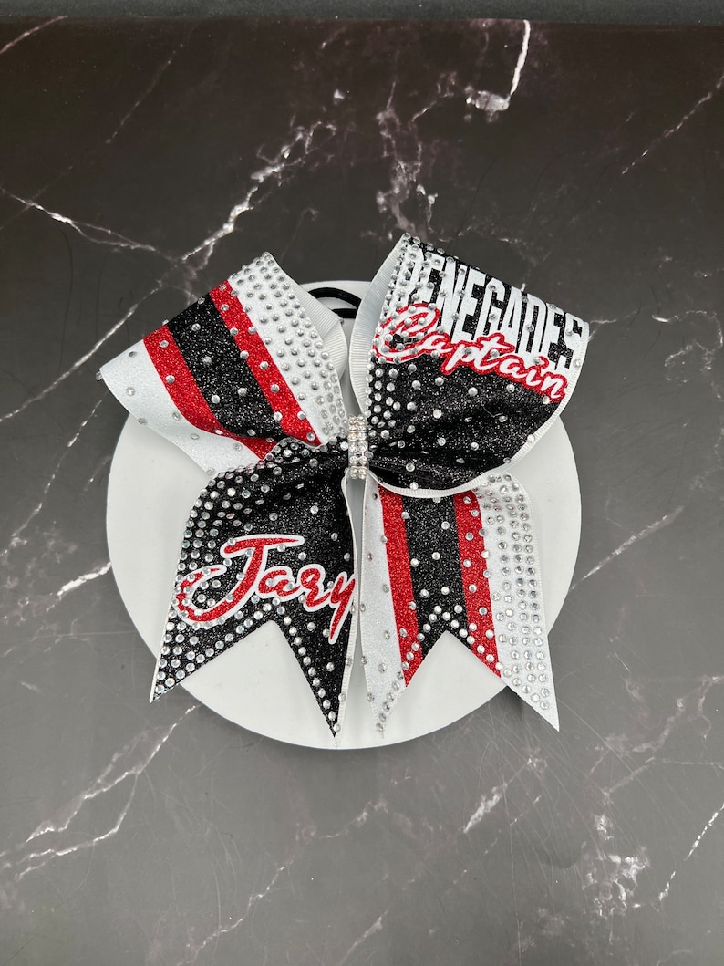 Op de afbeelding: Een cheerleading strik met een wit, zwart en rood ontwerp. De strik heeft de woorden "Renegades Captain" en "Jary" in rode en witte letters. De strik is versierd met strass-steentjes.