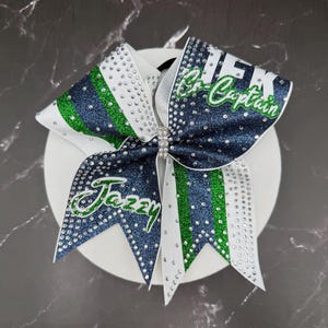 Op de afbeelding: Een cheerleading strik in wit, marineblauw en groen. De strik is versierd met strass en heeft de tekst "Jazzy" en "Co-Captain" in groene letters. In het midden van de strik zit een strass sluiting.