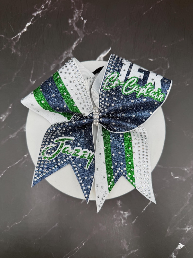 Op de afbeelding: Een cheerleading strik in wit, marineblauw en groen. De strik is versierd met strass en heeft de tekst "Jazzy" en "Co-Captain" in groene letters. In het midden van de strik zit een strass sluiting.