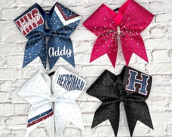 May include: Four glitter cheerleading bows in various colors. One is navy blue with "Mustangs" and "Addy" text, another is hot pink, a third is white with red and blue stripes and "Herriman" text, and the last is black with a blue "H".