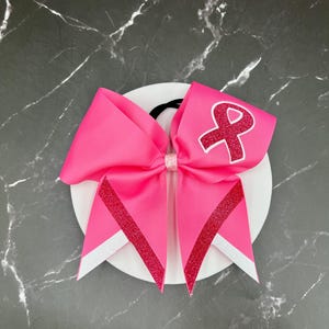 May include: A bright pink hair bow with a glittery red ribbon design and a white ribbon accent. The bow features a pink ribbon with a red ribbon symbol. The bow is attached to a black elastic band.