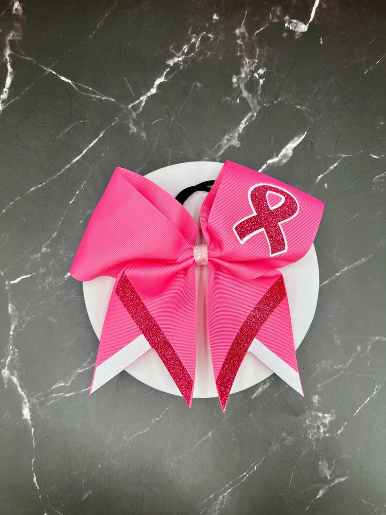 May include: A bright pink hair bow with a glittery red ribbon design and a white ribbon accent. The bow features a pink ribbon with a red ribbon symbol. The bow is attached to a black elastic band.