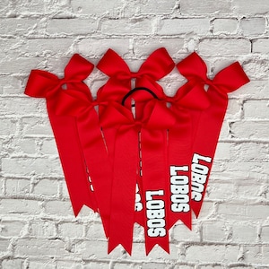 May include: Six red cheerleading bows with the word "LOBOS" printed in white block letters. Each bow has a large bow at the top and long, pointed ribbons. The bows are arranged against a white brick wall.