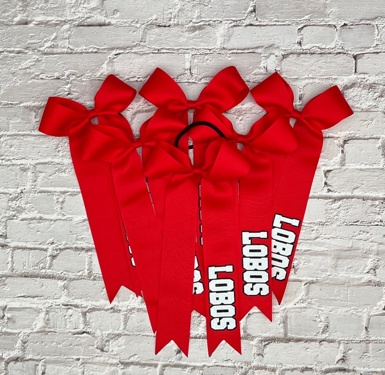 May include: Six red cheerleading bows with the word "LOBOS" printed in white block letters. Each bow has a large bow at the top and long, pointed ribbons. The bows are arranged against a white brick wall.