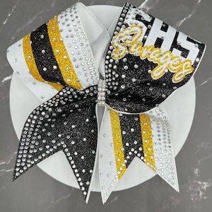 Op de afbeelding: Een grote cheerleading strik in wit, zwart en goud. De strik is versierd met strass-steentjes en heeft het woord "Savages" in goud. De strik ligt op een witte ondergrond. Geschikt voor cheerleading.