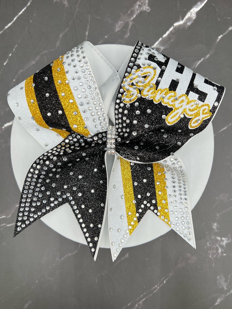 Op de afbeelding: Een grote cheerleading strik in wit, zwart en goud. De strik is versierd met strass-steentjes en heeft het woord "Savages" in goud. De strik ligt op een witte ondergrond. Geschikt voor cheerleading.