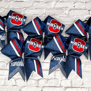 May include: Six navy blue glitter cheer bows with red and white accents. Each bow features the word "ROYALS" in red and white, and some have personalized names like "Camila", "Samantha", and "Kayla".