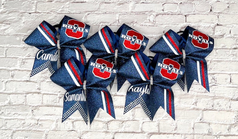 May include: Six navy blue glitter cheer bows with red and white accents. Each bow features the word "ROYALS" in red and white, and some have personalized names like "Camila", "Samantha", and "Kayla".