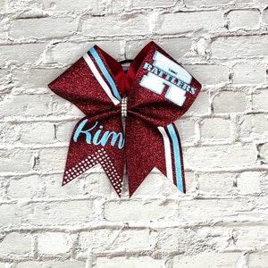 May include: A maroon glitter cheerleading bow with light blue and white accents. The bow features the name "Kim" in light blue script and the word "Rattlers" in white block letters. A rhinestone embellishment is at the center.