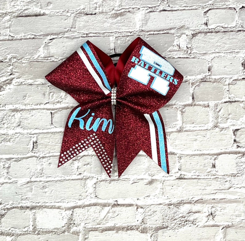 May include: A maroon glitter cheerleading bow with light blue and white accents. The bow features the name "Kim" in light blue script and the word "Rattlers" in white block letters. A rhinestone embellishment is at the center.