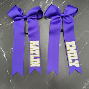 May include: Two purple cheerleading hair bows with the names "KAYLIN" and "EMILY" in gold glitter lettering. Each bow features a large bow at the top and long, ribbon tails.