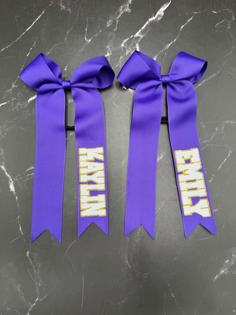 May include: Two purple cheerleading hair bows with the names "KAYLIN" and "EMILY" in gold glitter lettering. Each bow features a large bow at the top and long, ribbon tails.
