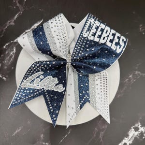 Op de afbeelding: Een cheerleading strik in marineblauw en wit, versierd met zilveren glitter en strass. De strik toont de naam "Jeebees" in witte blokletters en de naam "Lilla" in cursief.