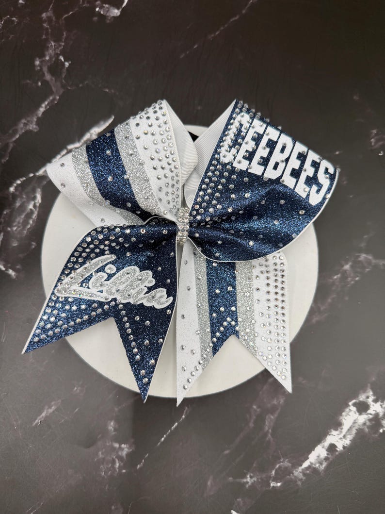 Op de afbeelding: Een cheerleading strik in marineblauw en wit, versierd met zilveren glitter en strass. De strik toont de naam "Jeebees" in witte blokletters en de naam "Lilla" in cursief.