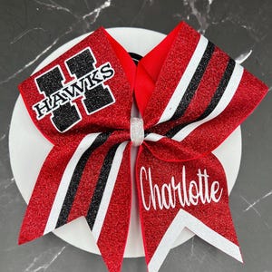 May include: A red glitter cheer bow featuring black and white striped accents. The left side displays a black and white HAWKS logo, while the right side features the name Charlotte in white script. A small silver glitter center knot secures the bow.