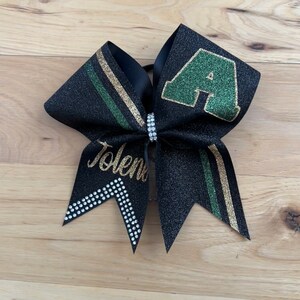 May include: Black glitter cheer bow with a green and gold stripe design. The bow features a green letter "A" and the name "Tolena" in gold script. A rhinestone band accents the center.