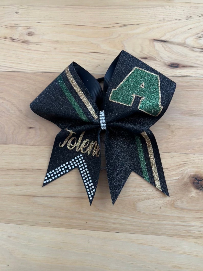 May include: Black glitter cheer bow with a green and gold stripe design. The bow features a green letter "A" and the name "Tolena" in gold script. A rhinestone band accents the center.