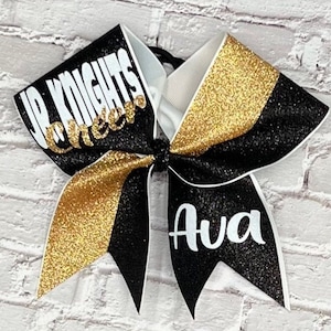 May include: A cheerleading bow with black, gold glitter, and white accents. The bow features the text "UP KNIGHTS Cheer" and the name "Ava" in white. The bow is likely for a school or team.