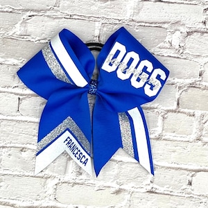 May include: A blue cheerleading bow with white and silver accents. The word "DOGS" is printed in white on the top of the bow. The name "FRANCESCA" is printed in white on the bottom of the bow.