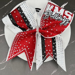 Op de afbeelding: Een rode, witte en zwarte cheerleading strik met de tekst "KMS Bulldogs". De strik is versierd met strass en heeft een glitterafwerking. De strik ligt op een wit oppervlak.