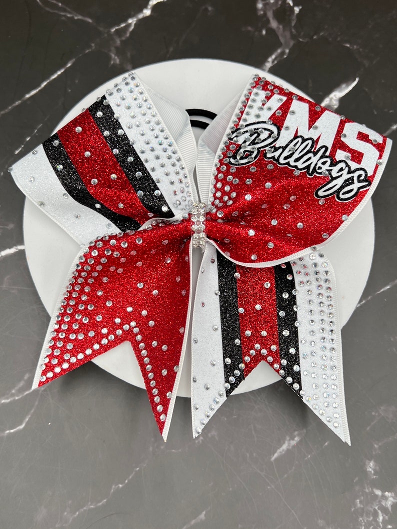 Op de afbeelding: Een rode, witte en zwarte cheerleading strik met de tekst "KMS Bulldogs". De strik is versierd met strass en heeft een glitterafwerking. De strik ligt op een wit oppervlak.