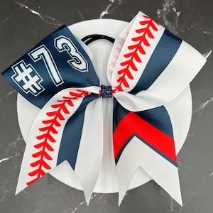 May include: A hair bow featuring a navy blue, white, and red color scheme with a baseball stitch pattern. The left side displays the text #73 in white with a navy blue outline. The center is secured with a dark blue glittery fabric knot.