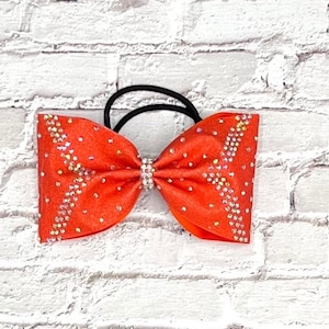 May include: A vibrant orange hair bow with sparkling rhinestones. The bow is centered with a rhinestone clasp and attached to a black elastic band. The bow is a bright, eye-catching accessory.