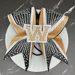 May include: A cheer bow featuring black glitter fabric, gold glitter accents, and white trim. The bow is embellished with numerous clear rhinestones and features a central white letter W with gold glitter fill and rhinestone detailing.
