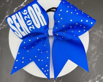 May include: A royal blue cheerleading bow with the word "SENIOR 2026" in white. The bow is adorned with small, sparkling rhinestones and has a silver ribbon accent at the center. The bow is attached to a black elastic band.