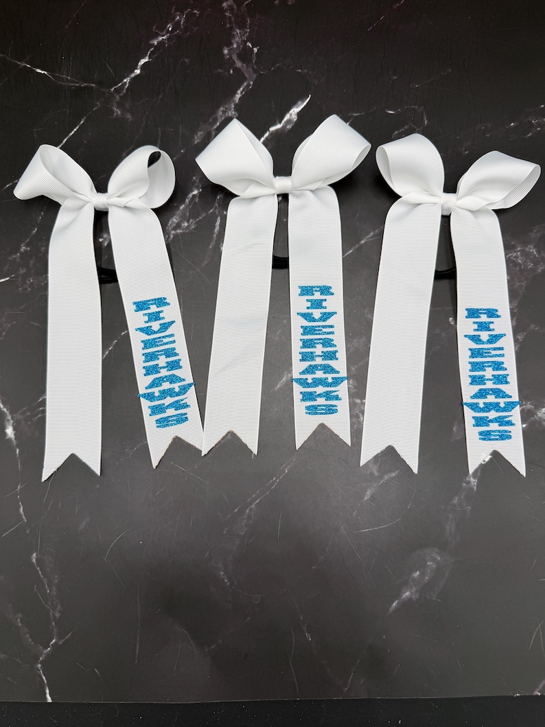 May include: Three white hair bows with long, ribbon tails. Each tail has the word "RIVERHAWKS" in blue glitter. The bows are on black elastic bands, suitable for ponytails or braids.