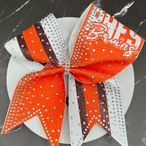 Op de afbeelding: Een grote oranje en witte cheerleading strik met de tekst "BHHS Bruins" in het wit. De strik heeft glitteraccenten, strass-steentjes en een centrale strass-versiering. De strik ligt op een wit oppervlak.