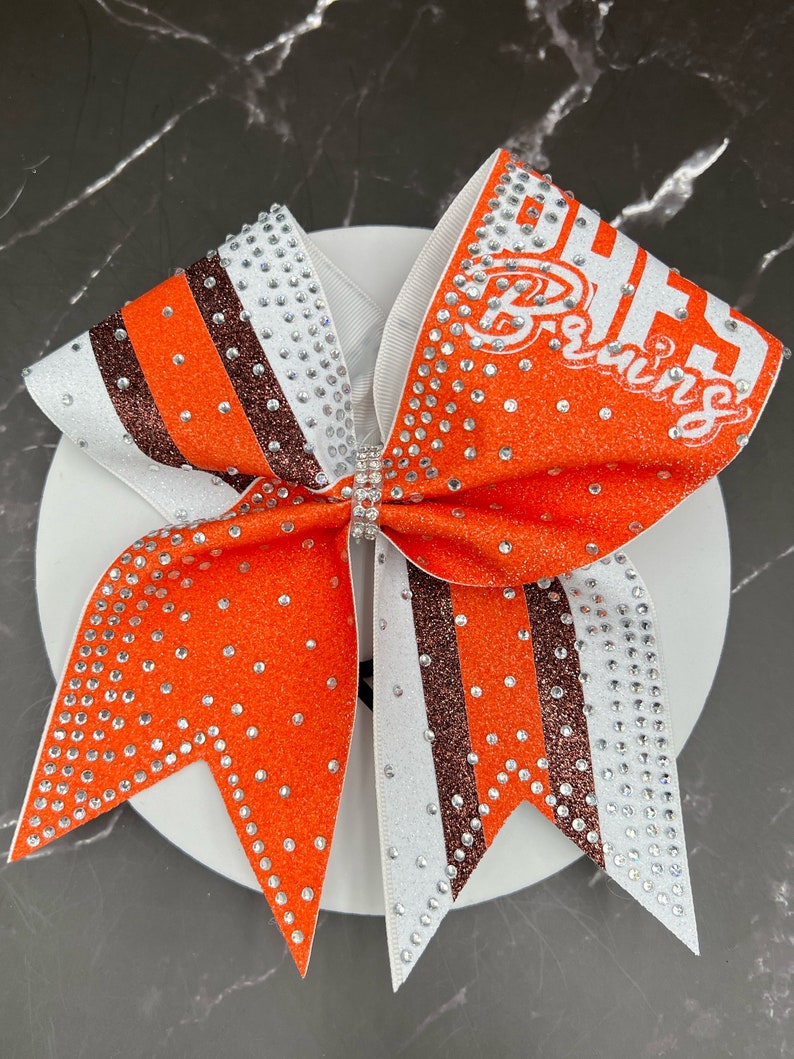 Op de afbeelding: Een grote oranje en witte cheerleading strik met de tekst "BHHS Bruins" in het wit. De strik heeft glitteraccenten, strass-steentjes en een centrale strass-versiering. De strik ligt op een wit oppervlak.