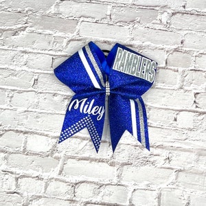 May include: A blue glitter cheerleading bow with white and silver accents. The bow features the name "Miley" and the word "Ramblers". Rhinestones and a silver stripe add detail to the bow.