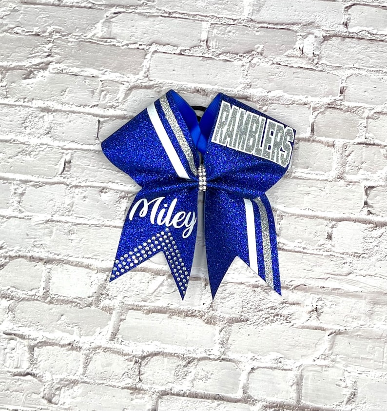 May include: A blue glitter cheerleading bow with white and silver accents. The bow features the name "Miley" and the word "Ramblers". Rhinestones and a silver stripe add detail to the bow.