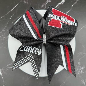 May include: A black glitter cheerleading bow with red and white stripes and the word "Lancers" in white script. The bow features the name "Palombi" in red block letters.