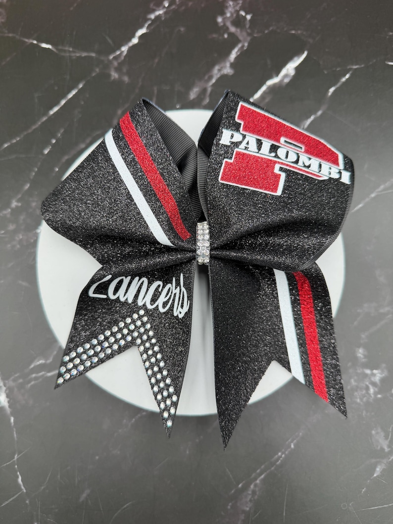 May include: A black glitter cheerleading bow with red and white stripes and the word "Lancers" in white script. The bow features the name "Palombi" in red block letters.