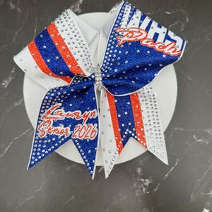 Op de afbeelding: Een cheerleading strik met een wit, blauw en rood ontwerp. De strik heeft de tekst "Dash" en "Kamryn Senior 2026" in rood en wit. De strik is versierd met strass.