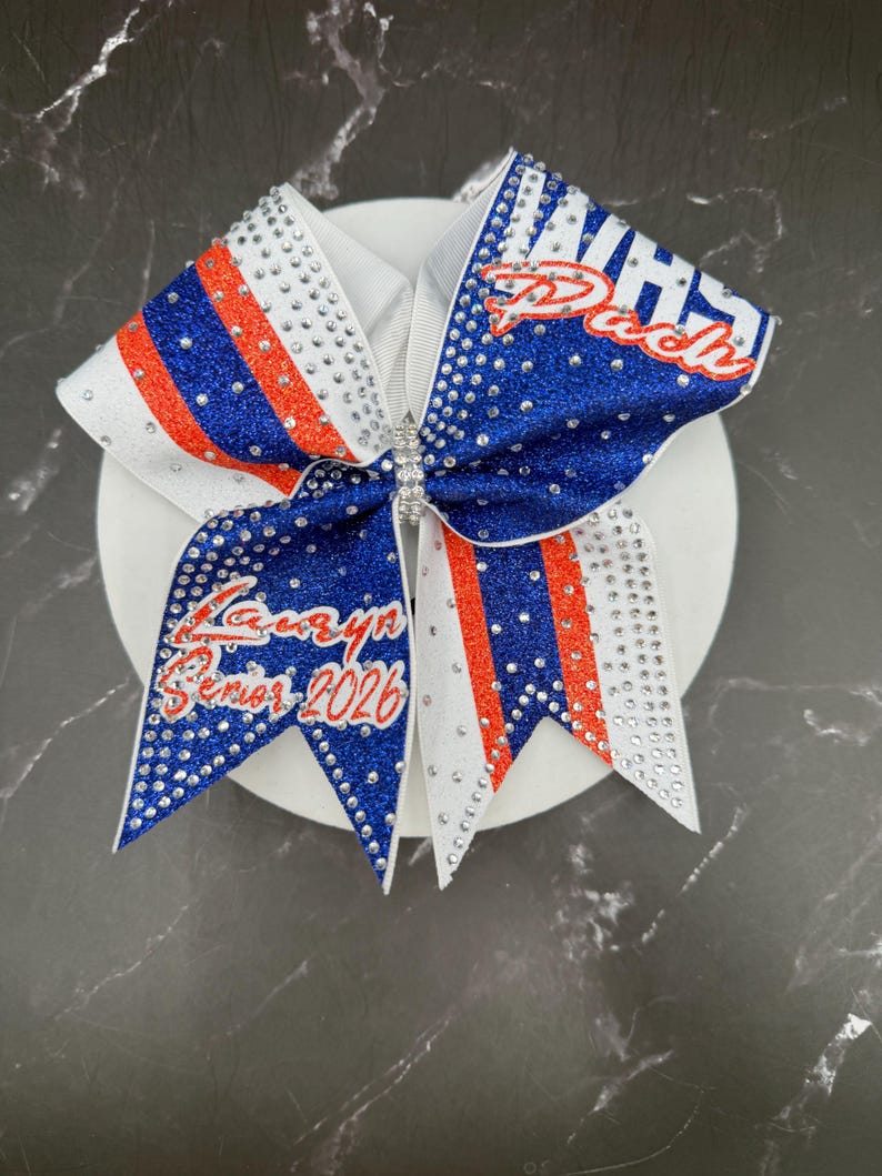 Op de afbeelding: Een cheerleading strik met een wit, blauw en rood ontwerp. De strik heeft de tekst "Dash" en "Kamryn Senior 2026" in rood en wit. De strik is versierd met strass.