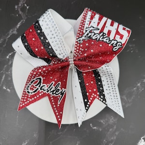 Op de afbeelding: Een cheerleading strik met rode glitter, zwarte en witte strepen. De strik heeft de tekst "VHS Indians" en "Oakley" in witte letters en is versierd met strass.