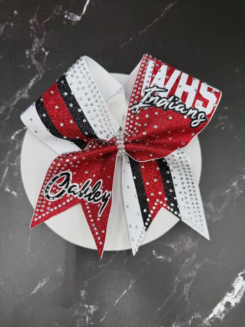 Op de afbeelding: Een cheerleading strik met rode glitter, zwarte en witte strepen. De strik heeft de tekst "VHS Indians" en "Oakley" in witte letters en is versierd met strass.